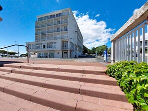 A multi-story building with a flat roof and balconies, a pedestrian crossing, and a staircase leading up to the entrance.