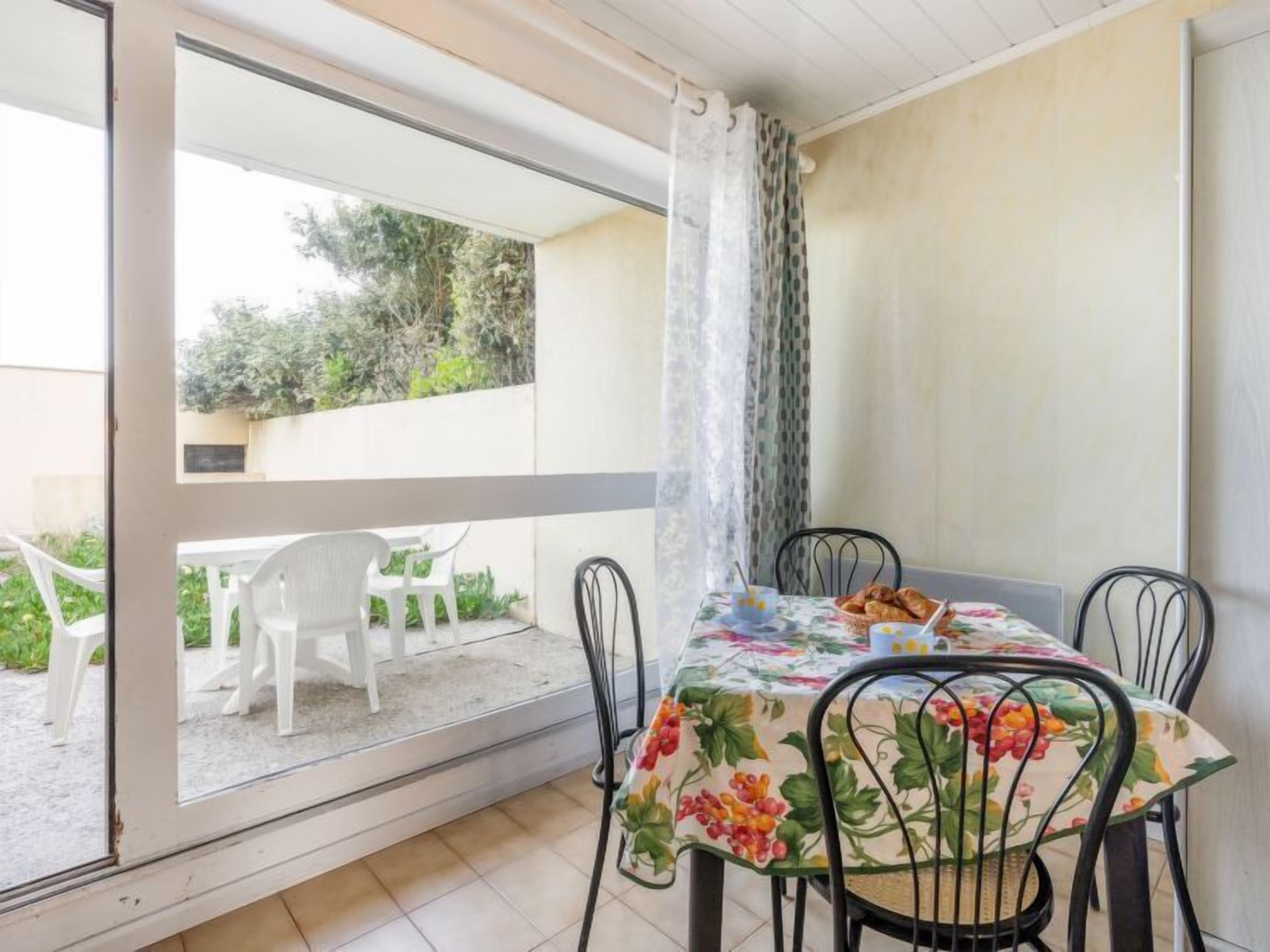 A dining area with a table set for two, a view of the garden through a glass door, and a white chair outside.