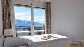 A hotel room with two beds, a window with a view of mountains, and a lamp on the left. - Jeanne D'Arc Apt. D-33 by Interhome (Crans-Montana)