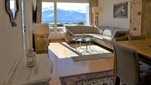A living room with a sofa, a round coffee table, a dining table, and a view of mountains. - Jeanne D'Arc Apt. D-33 by Interhome (Crans-Montana)