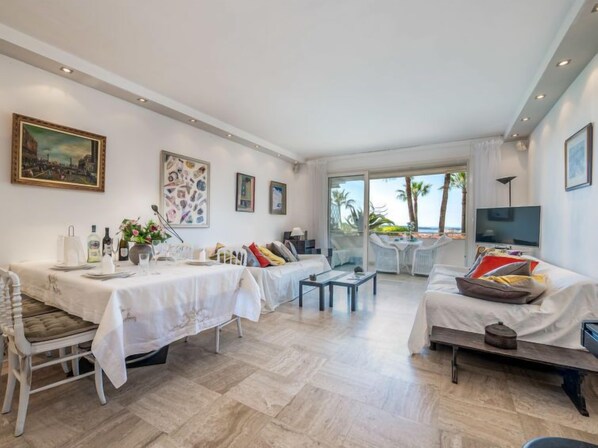 A spacious living area with a dining table set for two, a sofa, and a balcony with a view of palm trees.