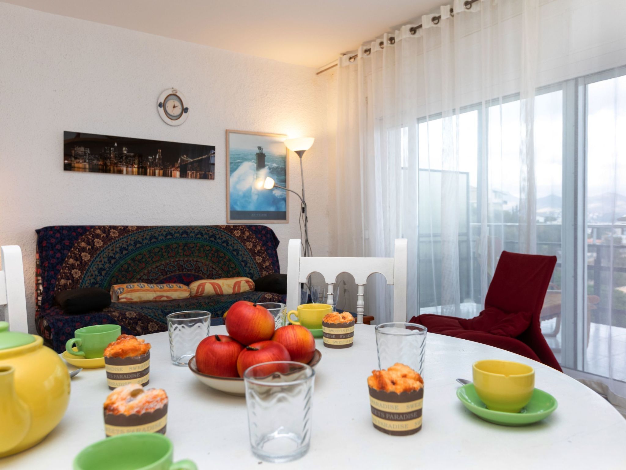 A dining area with a table set for a meal, a sofa with a patterned throw, a cityscape view through large windows, and a clock on the wall.