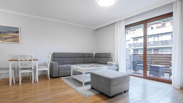 Ett modernt vardagsrum med en grå soffa, ett vitt soffbord och en tillhörande pall. Det finns en matplats med vita stolar och ett bord. Rummet har trägolv och en stor skjutdörr i glas som leder ut till en balkong med möbler i rotting.