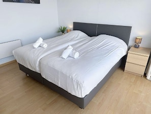 A double bed with white linens, a dark headboard, a bedside table with a lamp, and a small plant.