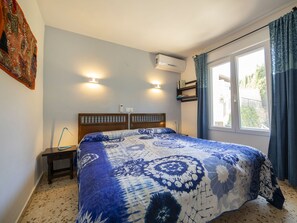 A bedroom with a bed, a bedside table, a wall-mounted air conditioner, a window with curtains, and a wall-mounted light fixture.