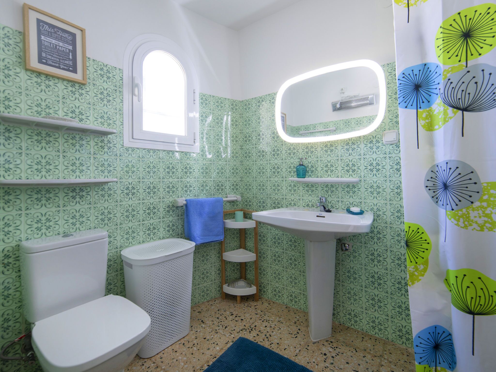 A bathroom with green patterned tiles, a white sink, a mirror, a window, and a blue towel.