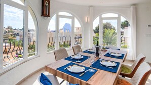 Un coin repas avec une table en bois dressée pour six personnes, de grandes fenêtres en arc de cercle et une vue sur un quartier résidentiel.