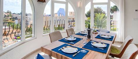 A dining area with a wooden table set for six, large arched windows, and a view of a residential area.