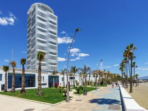 Un hôtel moderne à la façade incurvée, doté d’un jardin bien entretenu et d’une promenade en bord de mer bordée de palmiers.