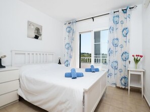 Une chambre avec un lit blanc, des serviettes bleues, une table de chevet, une petite table sur laquelle est posé un vase de fleurs, et une fenêtre ornée de rideaux à motifs floraux bleus.
