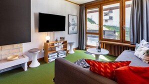 A modern living room with a flat-screen TV, a sofa, a small table, and a window with curtains. - Le Pramecou (Val Claret) by Interhome (Tignes)