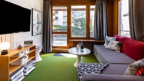 A modern living room with a sofa, a TV, a small table with a bottle, and a view of buildings through large windows. - Le Pramecou (Val Claret) by Interhome (Tignes)