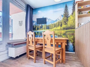A dining area with a wooden table and chairs, a TV mounted on the wall, and a scenic mural of a lake and mountains. - Névés 150 by Interhome (Val Thorens)