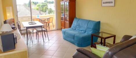 Een woonkamer met een blauwe bank, een bruine leren fauteuil, een glazen salontafel en een eethoek met een tafel en stoelen.