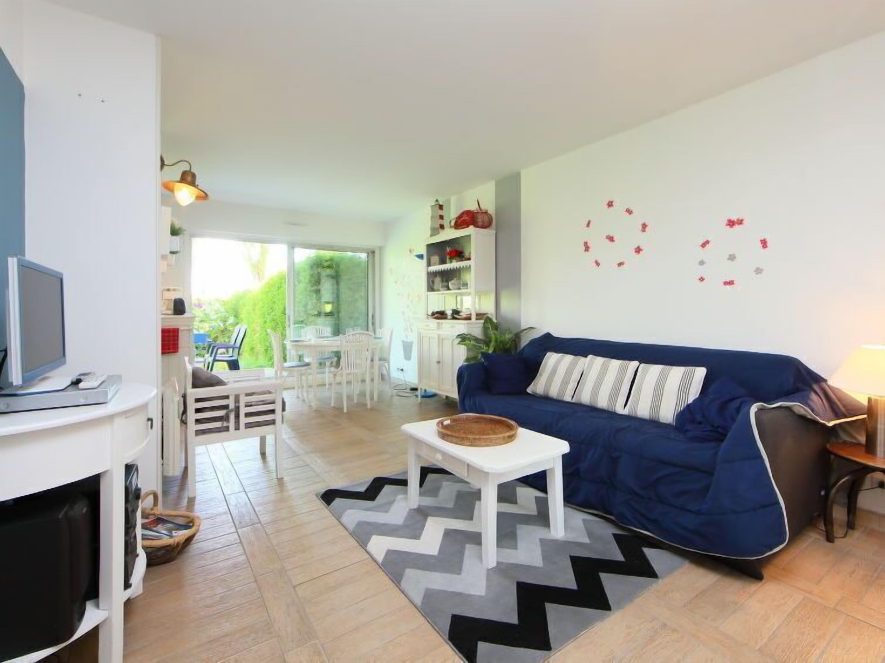 A modern living room with a blue sofa, a white coffee table, and a television on a white stand. There is a dining area with white chairs and a table, and a kitchen area with white cabinets and a red pot on the counter.