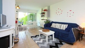 A modern living room with a blue sofa, a white coffee table, and a television on a white stand. There is a dining area with white chairs and a table, and a kitchen area with white cabinets and a red pot on the counter. - Le Garden by Interhome (Cabourg)