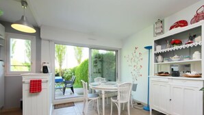 A bright kitchen with white cabinets, a dining table with chairs, and a view of the garden through the glass door. - Le Garden by Interhome (Cabourg)