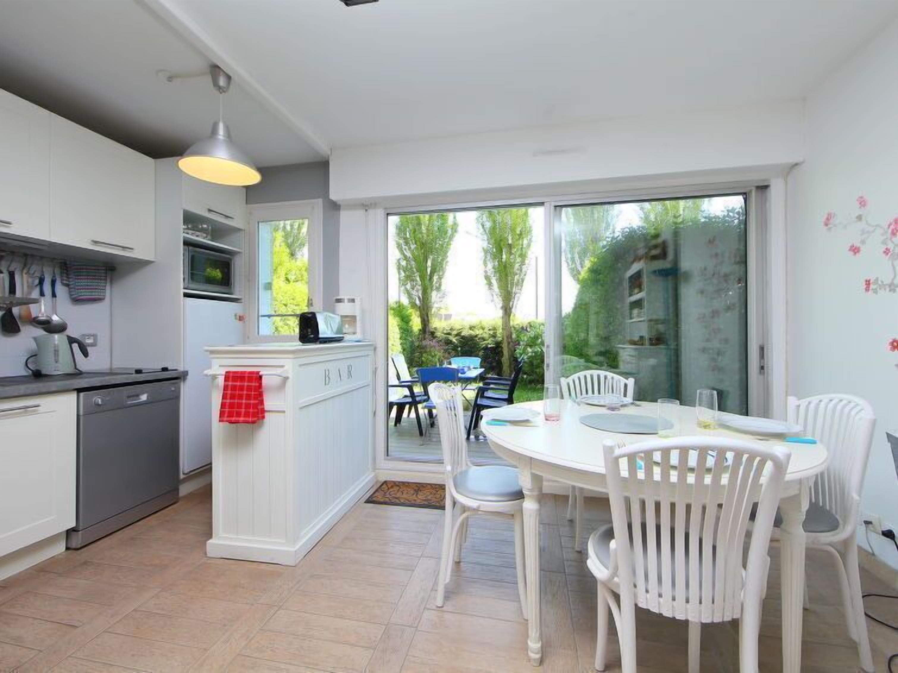 A modern kitchen with white cabinetry, a dining area with white chairs, and a view of the garden through large sliding glass doors.