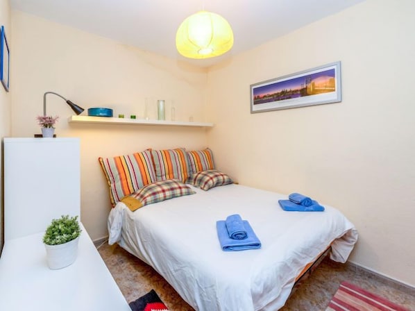 Un dormitorio con cama, almohadas, mesita de noche, lámpara, estante con objetos decorativos, planta en maceta, cuadro en la pared y lámpara colgante.
