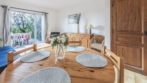 A cozy room with a wooden dining table, chairs, a large wooden door, and a view of a garden through a sliding glass door. - Bonporteau by Interhome (Cavalaire)