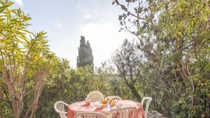 A round table with a red tablecloth and white chairs in a garden setting, surrounded by lush greenery and a stone pathway. - Bonporteau by Interhome (Cavalaire)