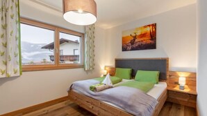 Quarto com cama de madeira, almofadas verdes, pintura de pôr do sol, janela com cortinas e luminária pendente.