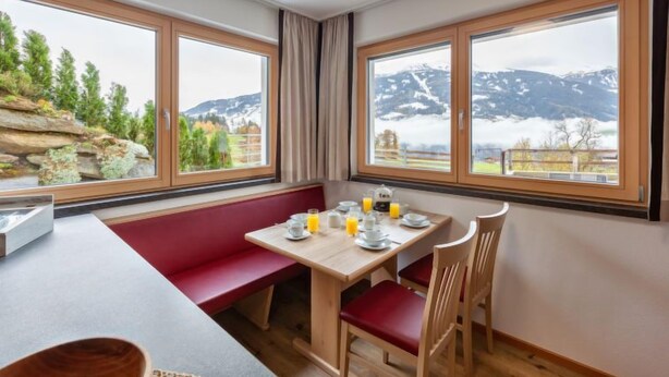 Uma área de refeições com mesa posta para o café da manhã, grandes janelas com vista para as montanhas e um banco vermelho.