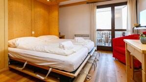 Una habitación con dos camas, un sofá rojo, una mesita con un jarrón y un balcón con sillas.