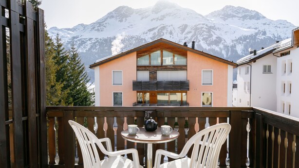 A balcony with two white plastic chairs and a small round table, overlooking a snow-covered mountain and a building with a peach facade. - Apartment Apt.17 by Interhome (Silvaplana-Surlej)