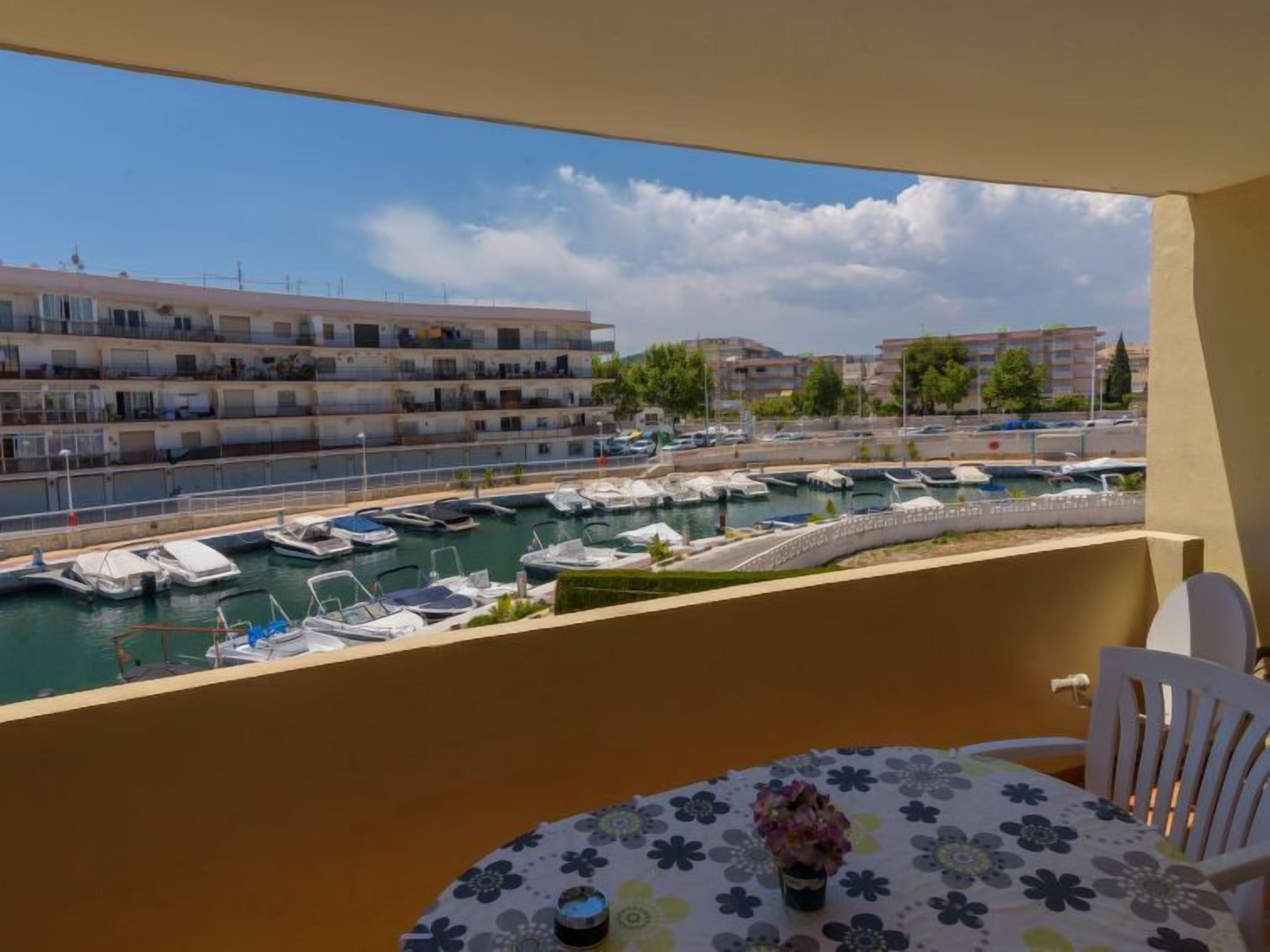 Un balcon donnant sur un port de plaisance, une table recouverte d’une nappe à motifs floraux et des chaises blanches.