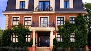 Una casa a due piani in mattoni con tetto scuro, un ingresso centrale con porta nera e un balcone con ringhiere in metallo decorate.