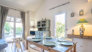 Uma área de refeições com mesa de madeira posta para quatro pessoas, cadeiras brancas, pequena cozinha com armários e uma janela com cortinas.