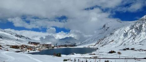 A snow-covered landscape with a lake, mountains, and buildings in the background.