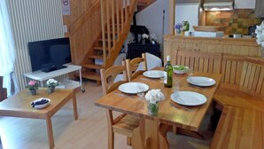 Un comedor de madera con una mesa dispuesta para cuatro comensales, un televisor sobre un mueble bajo y una escalera que sube a un nivel superior.