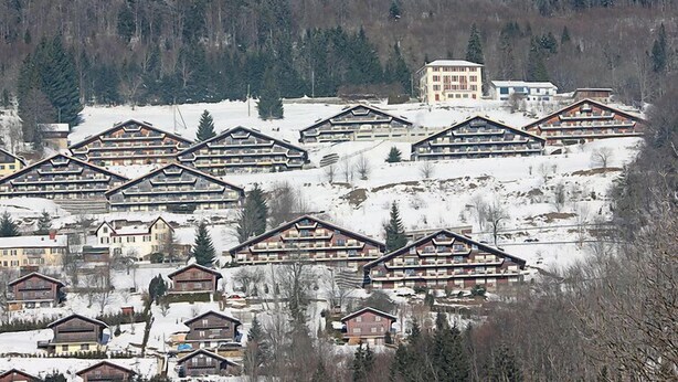 Un complejo turístico invernal cubierto de nieve con edificios de estilo chalet, un edificio central con techo rojo y un fondo de bosque.