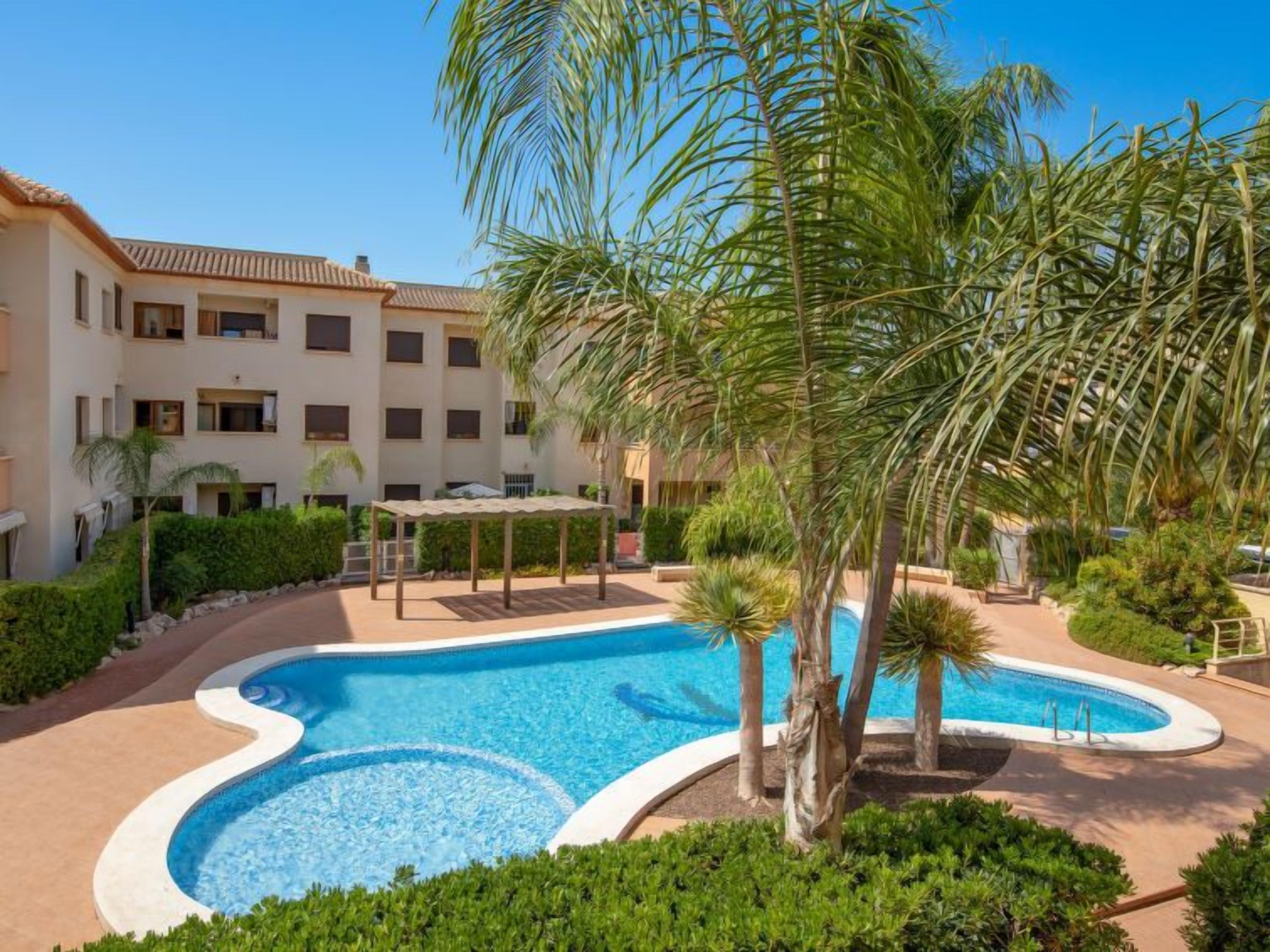 A swimming pool surrounded by palm trees and a multi-story building with balconies.
