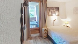 Une chambre à coucher comprenant un lit, une table de chevet, une lampe, un luminaire au plafond, une fenêtre avec des rideaux et une vue sur un espace extérieur.