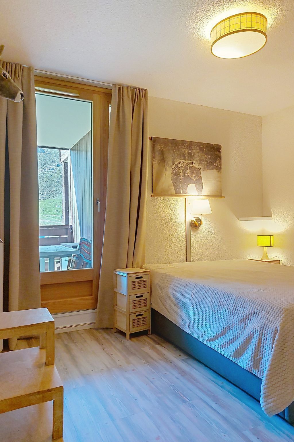 A bedroom with a bed, a desk, a chair, a window with curtains, and a wall-mounted lamp.