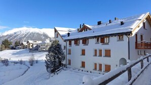 Un bâtiment enneigé, doté de nombreuses fenêtres et d’un balcon, entouré d’un paysage hivernal et avec des montagnes en arrière-plan.
