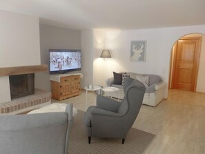 Una sala de estar con chimenea, televisor, un sillón gris, sofá, una mesita redonda y una lámpara.