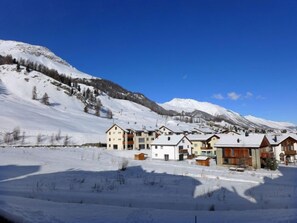 Un pueblo cubierto de nieve con edificios modernos, una pista de esquí y montañas al fondo.