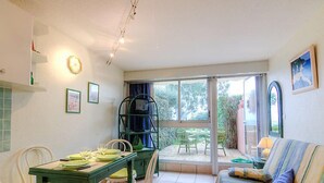 Quarto com mesa de jantar posta para quatro, uma cadeira verde, sofá com almofadas e uma grande janela com vista para o exterior.