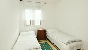 A small, clean room with two single beds, a wooden nightstand, a window with sheer curtains, and a patterned rug. - Apricot by Interhome (Balatonfenyves)