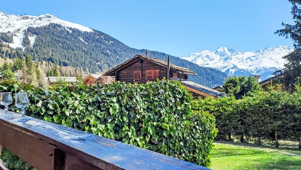 Eine Holzhütte vor einer Bergkulisse, umgeben von viel Grün und blauem Himmel.