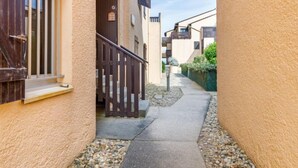 Une ruelle étroite entre deux bâtiments, avec une porte en bois, un chemin en béton et un petit espace aménagé en jardin.