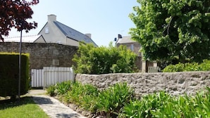 Un chemin de pierres mène à un bâtiment en pierre, entouré d’une clôture blanche et bordé d’une végétation luxuriante et d’arbres.
