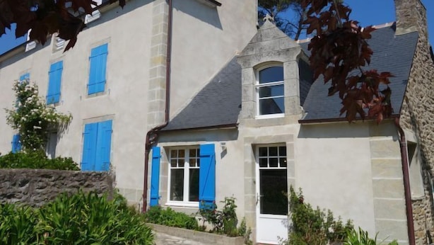 Un bâtiment à deux étages, doté d’une cheminée en pierre, d’une petite tour avec une fenêtre et d’une porte blanche.