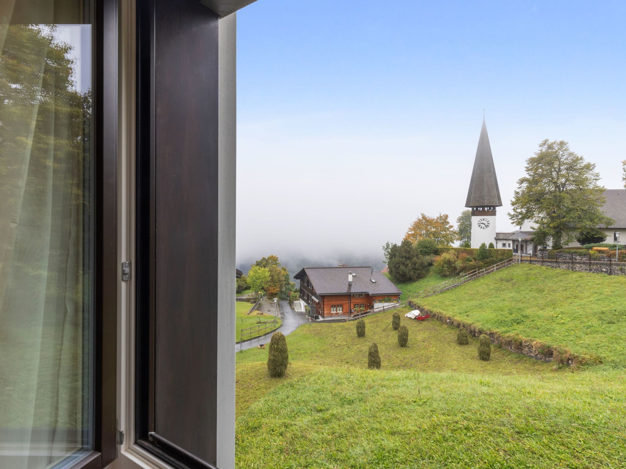 Une vue depuis une fenêtre donnant sur une église avec un clocher, un bâtiment rouge et une colline verdoyante.