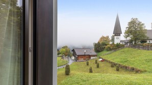 Vista da janela com igreja e campanário, um edifício vermelho e uma colina gramada.