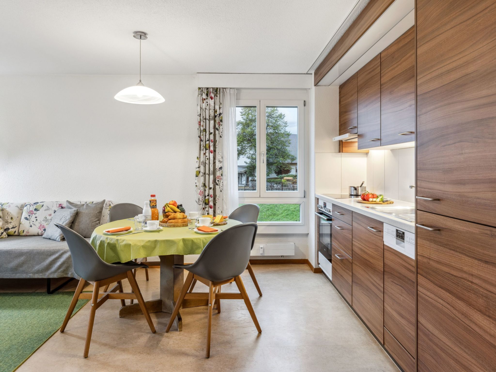 Une cuisine moderne avec des meubles en bois, une table à manger avec des chaises et une vue sur l’extérieur par une fenêtre.